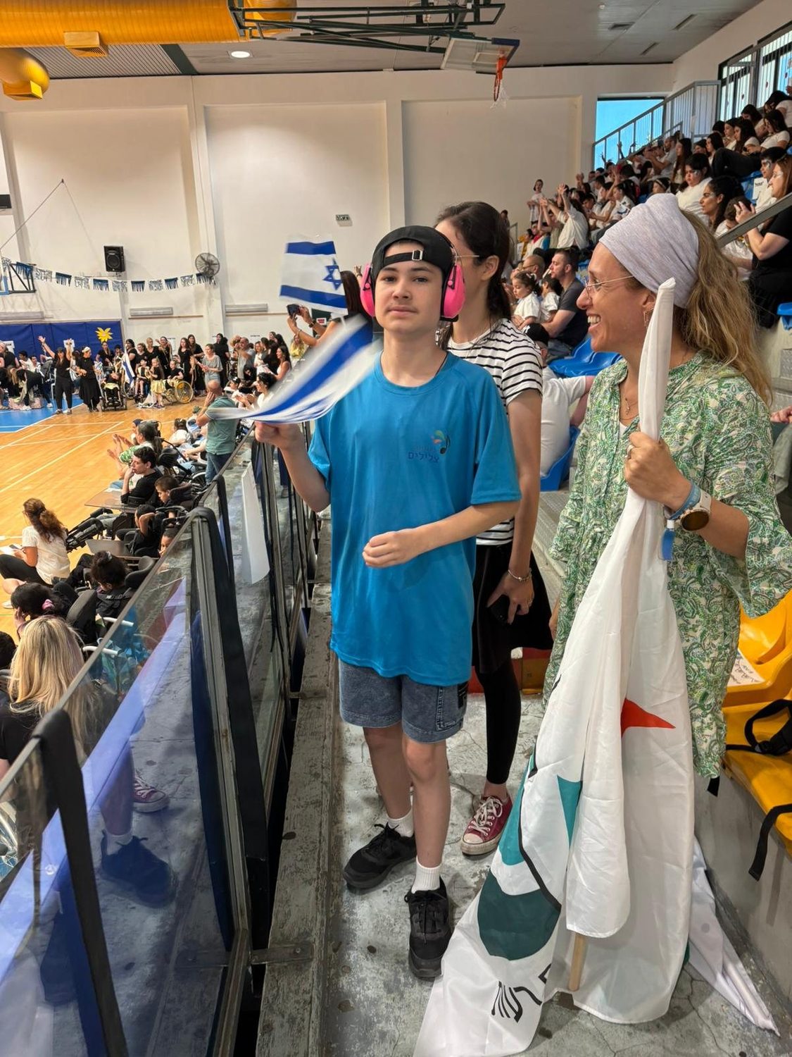 A teenage boy wearing noise-cancelling headphones holds a flag at the conference, with an aide beside him
